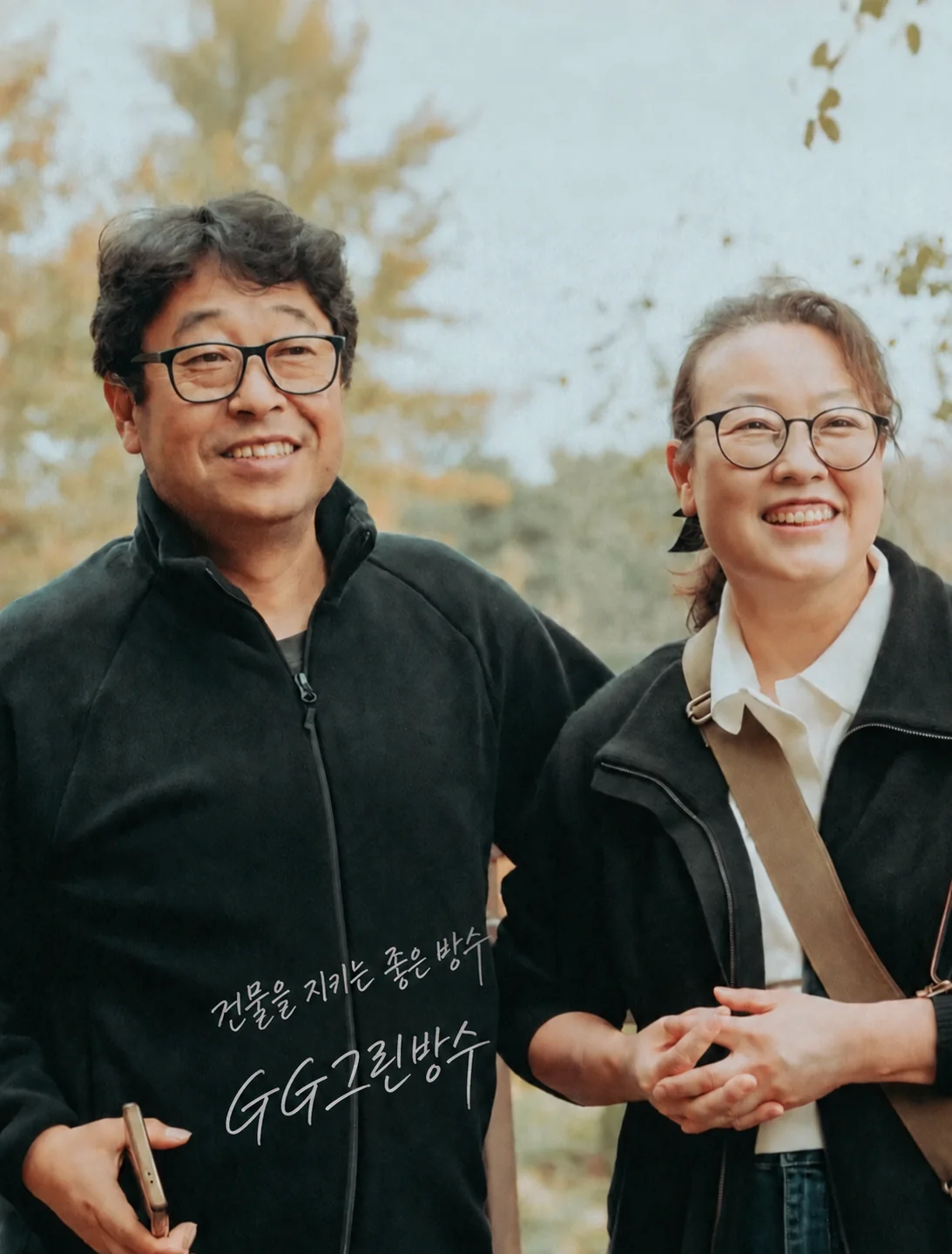 가을 야외에서 함께 미소 짓는 GG그린방수 두 구성원과 ‘건물을 지키는 좋은 방수 · GG그린방수’ 서명